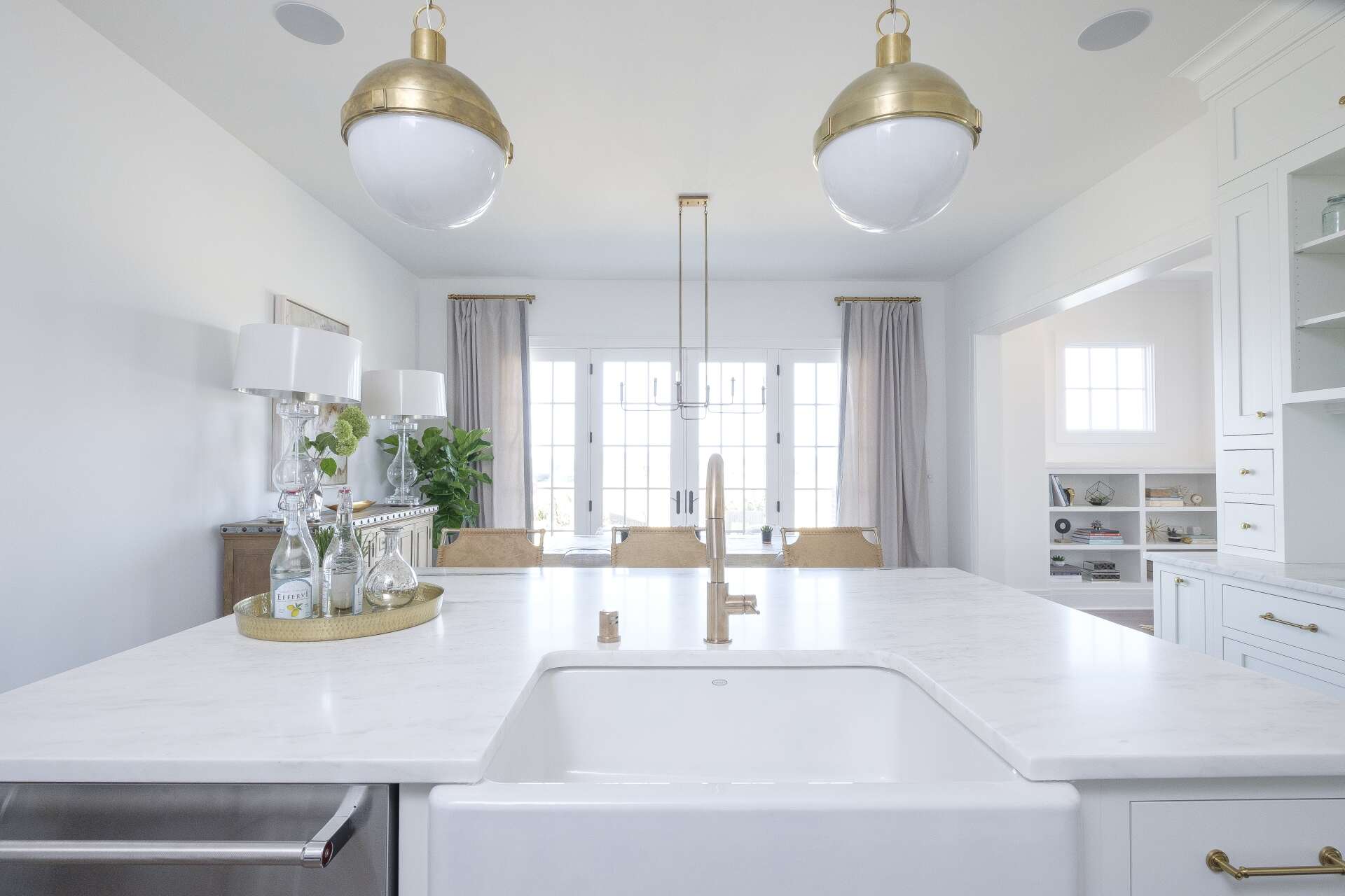 Bright white kitchen with gold pendant lights, large island, and dining area by sunlit windows.