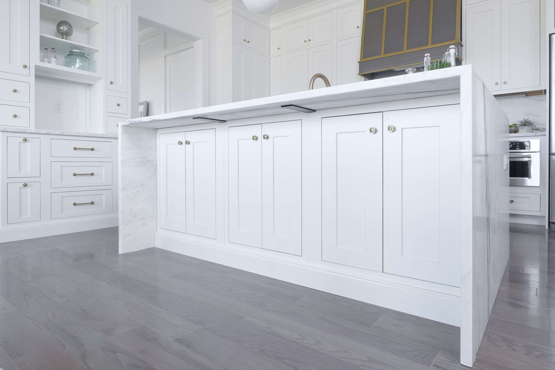 Bright white kitchen with an island, paneled cabinets, gray floor, and gold-accented range hood.