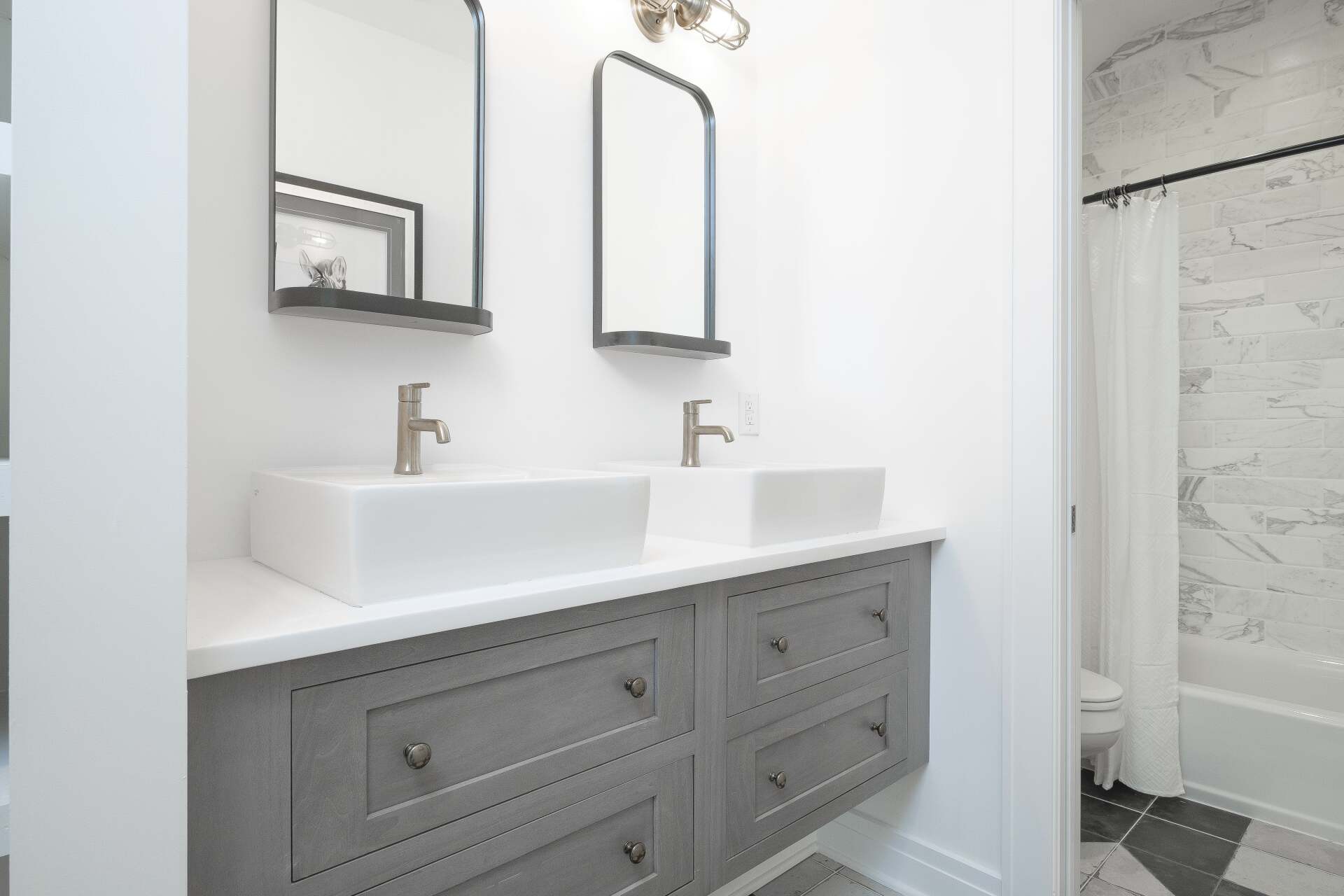 Modern white bathroom vanity with double sinks, gray cabinets, mirror, and shower visible in background