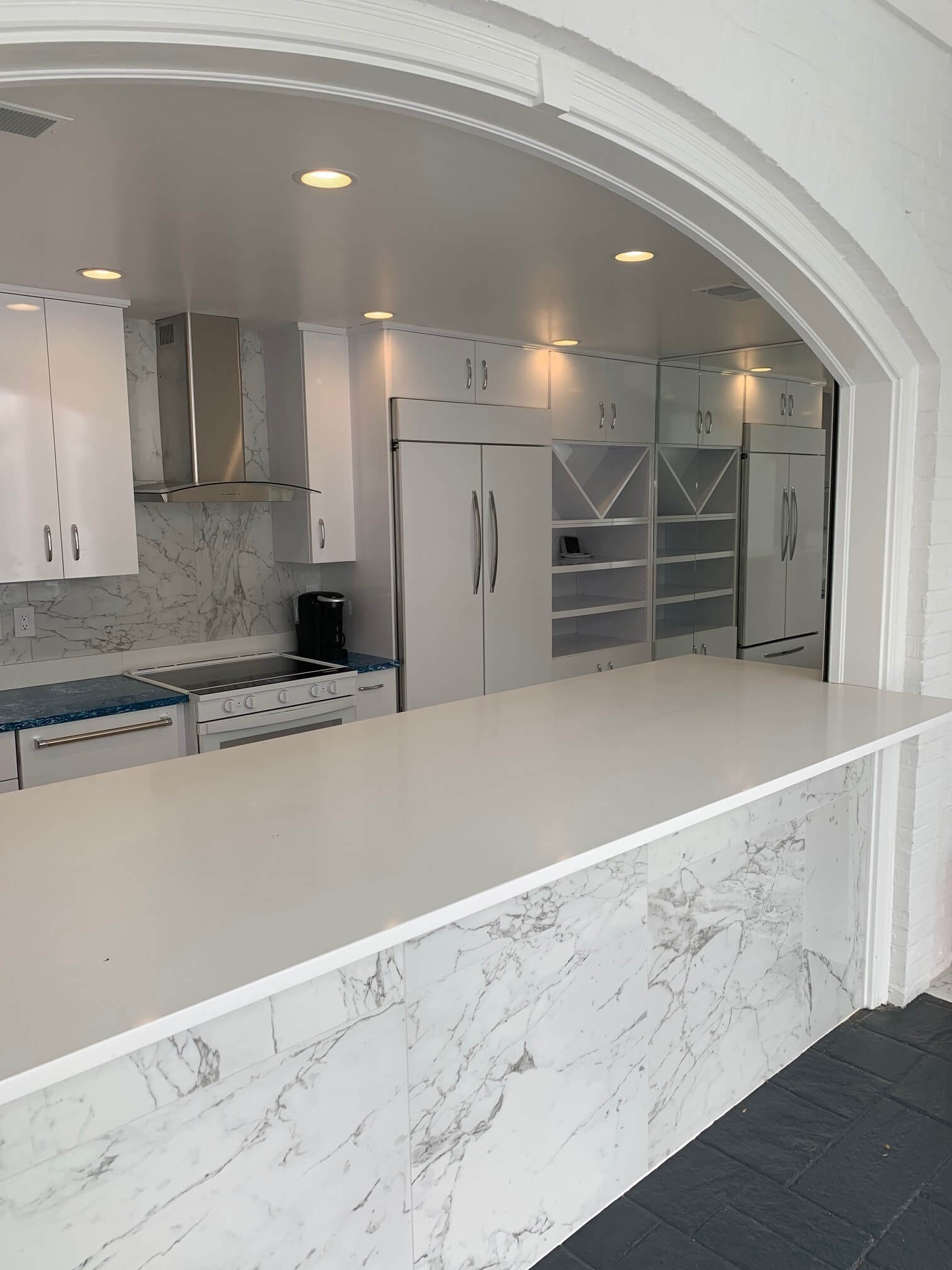 Bright white kitchen with marble island, arched pass-through, stainless appliances, and gray tiled floor
