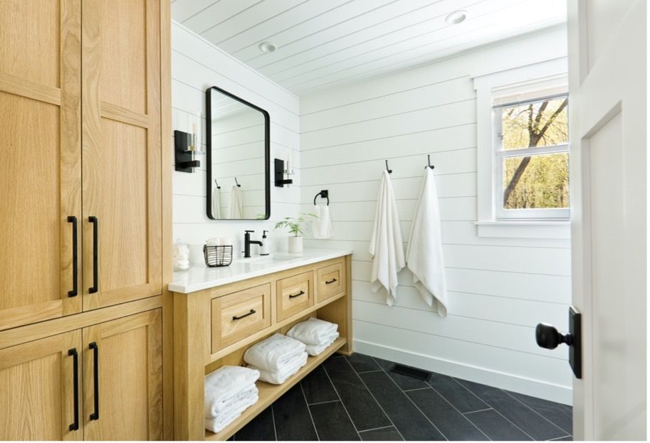 Bright bathroom with wood vanity, black tile floor, white shiplap walls, mirror, towels, and window.