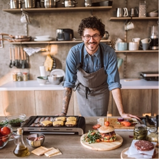 Chef smiling behind a kitchen counter with grilled food, fresh ingredients, and serving platters.