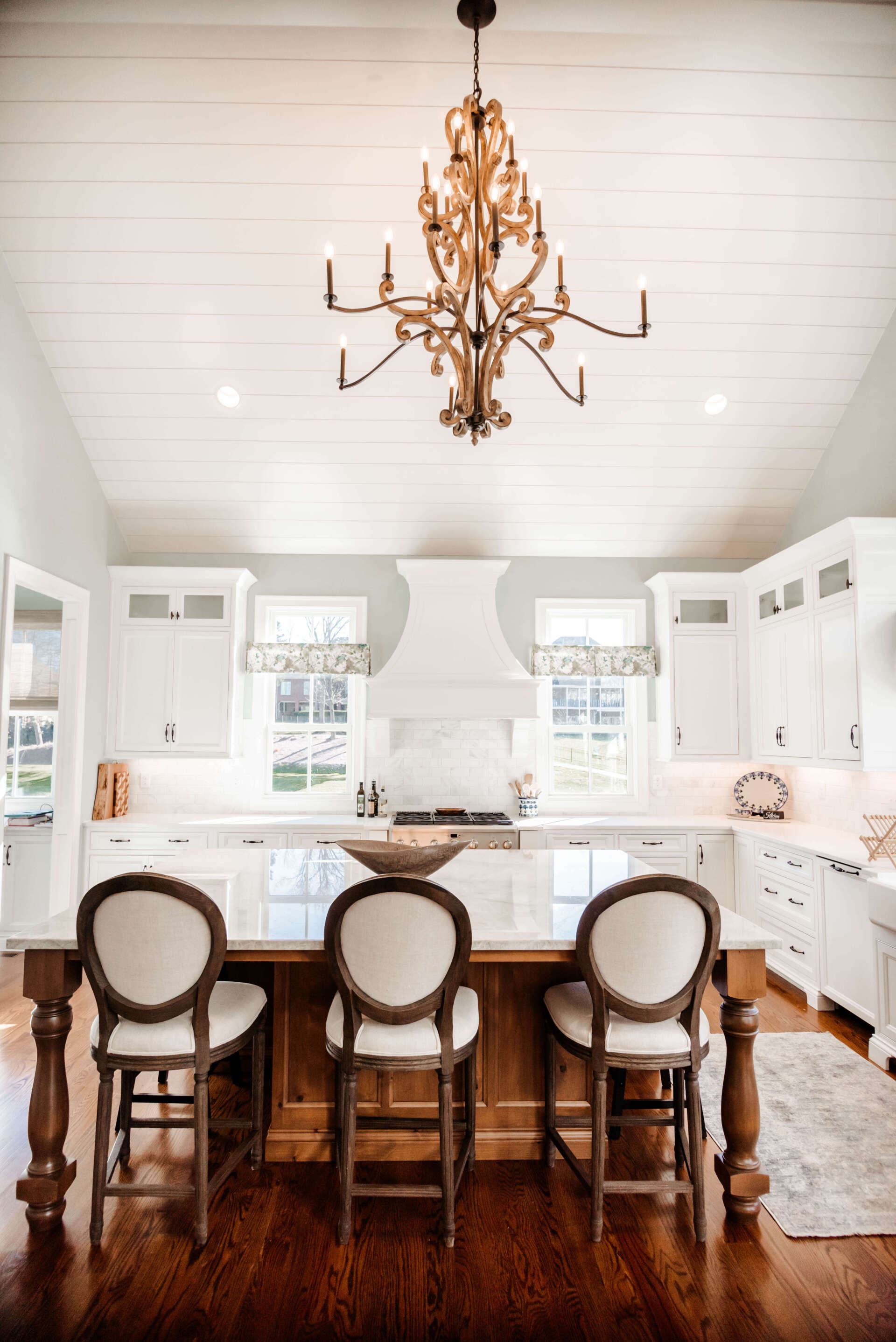Bright white kitchen with a large wooden island, bar stools, and a gold chandelier under a vaulted ceiling