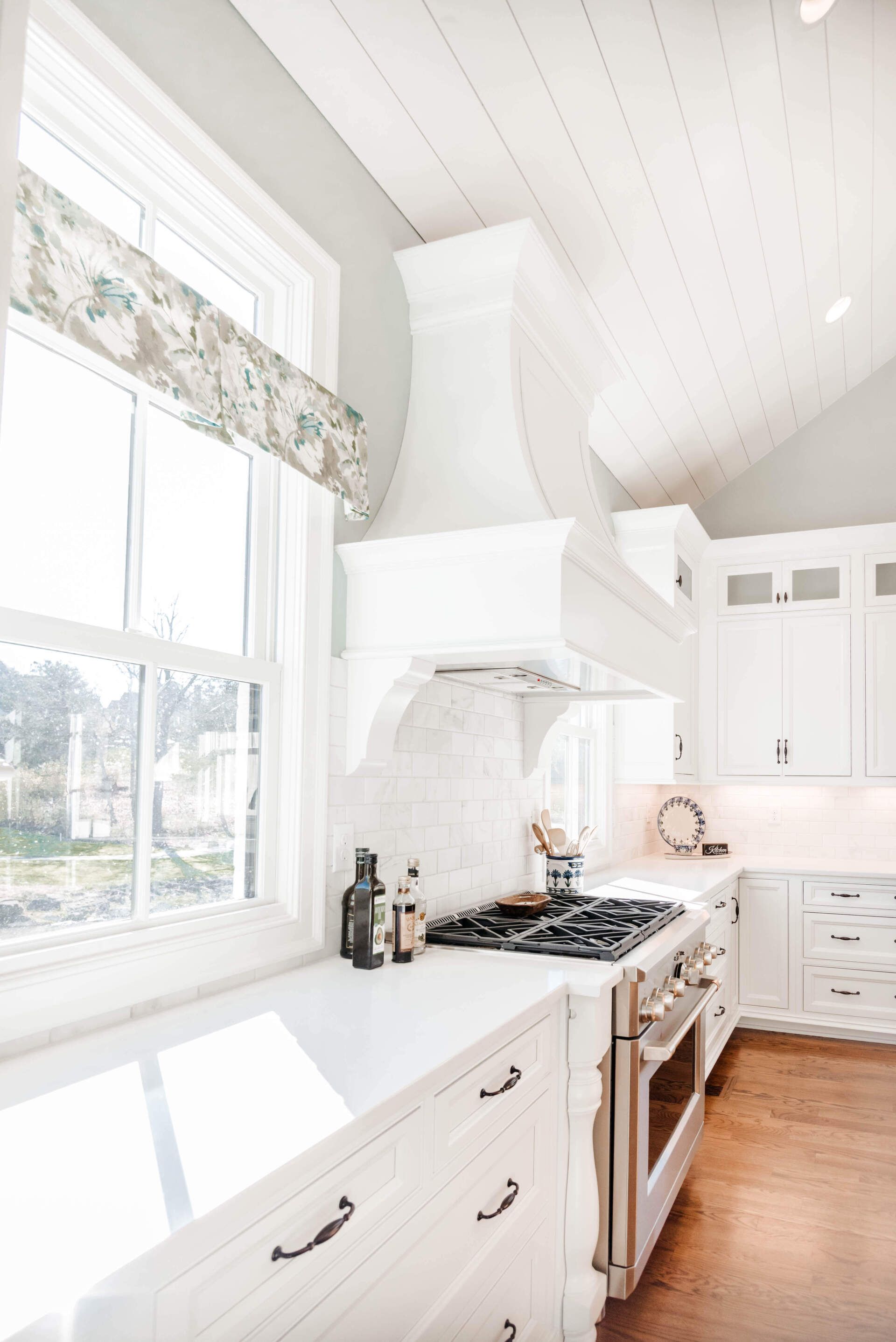Bright white kitchen with farmhouse hood, gas stove, cabinets, and sunlight from large windows