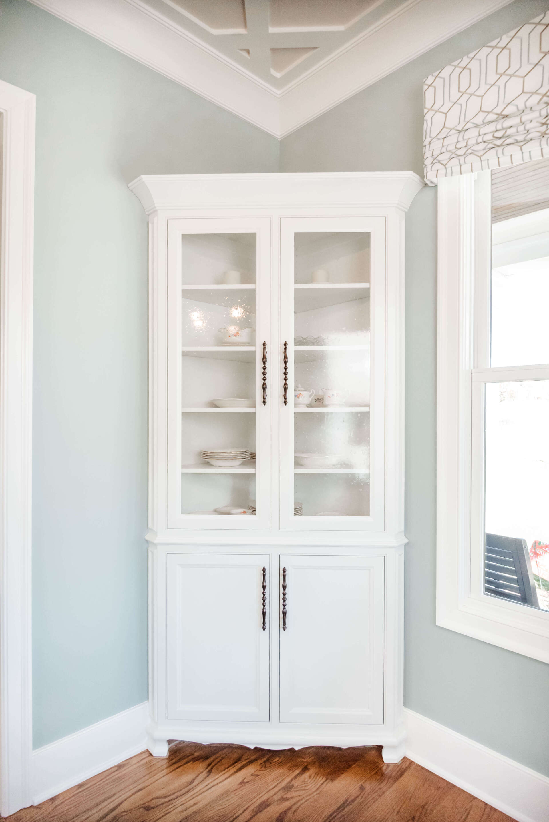 White corner china cabinet in a pale blue room with wood floor and window