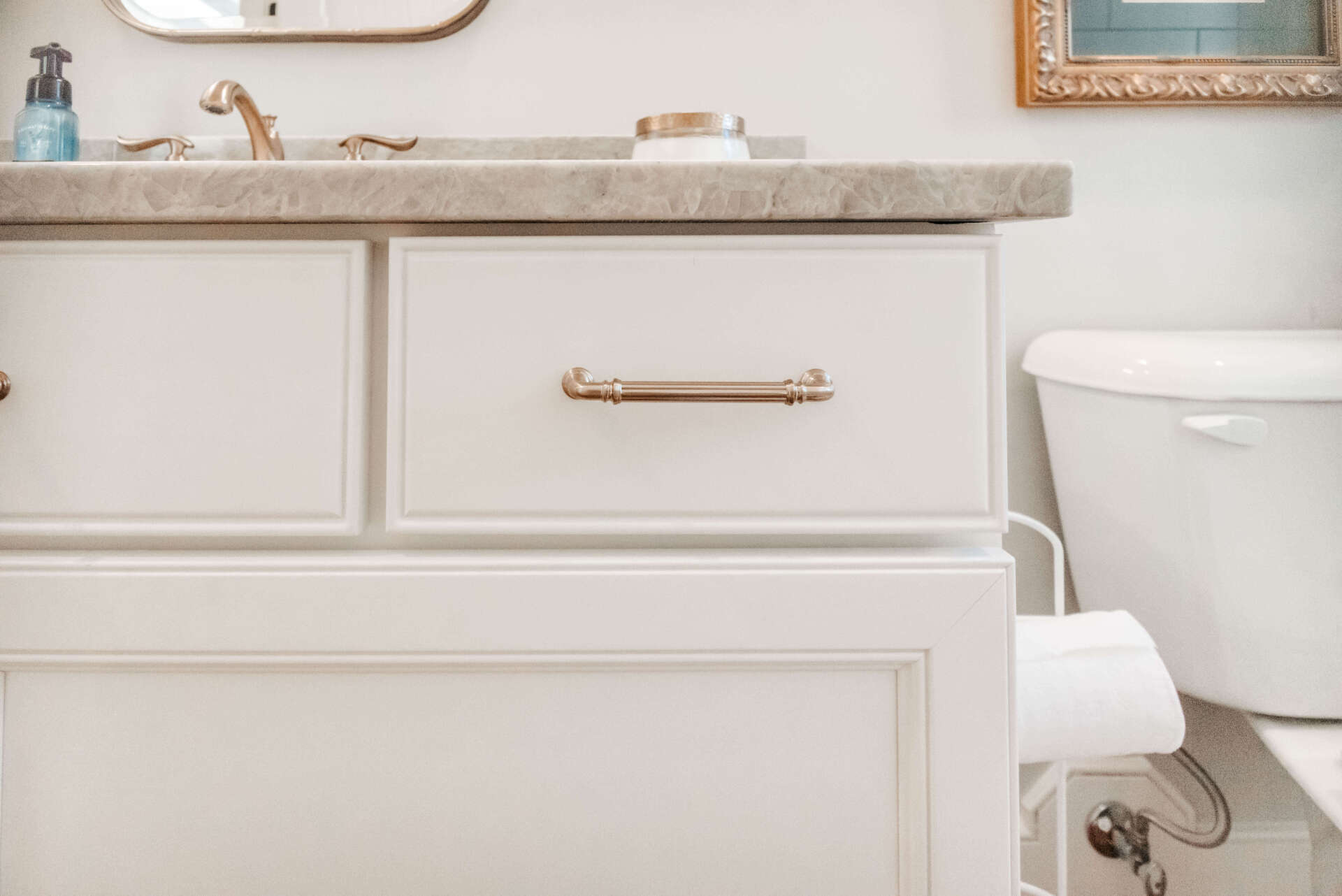 White bathroom vanity with brass handle, countertop sink, mirror, and toilet in a bright, neutral bathroom