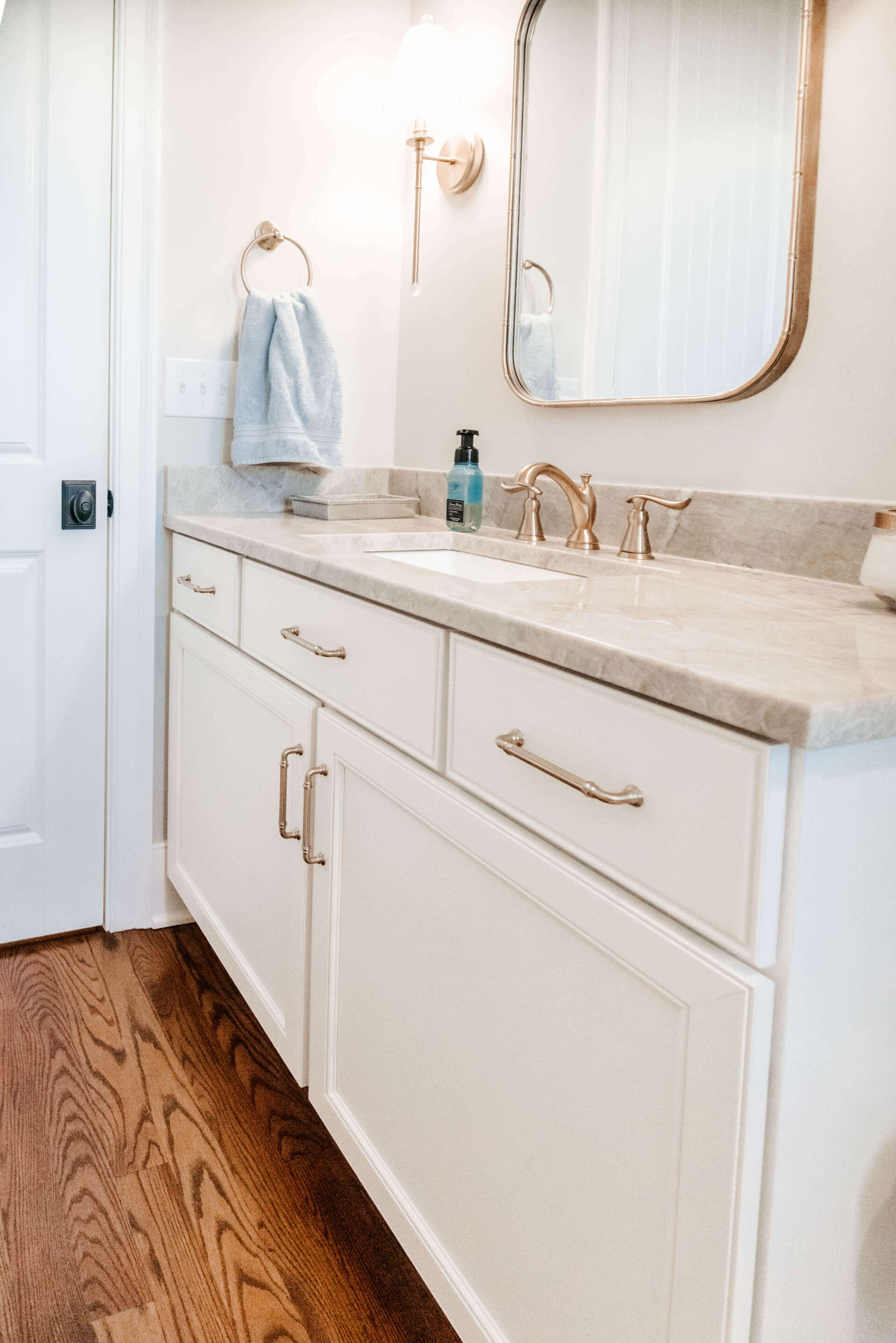 Bright white bathroom vanity with gold accents, marble countertop, and large mirror over wood floor.