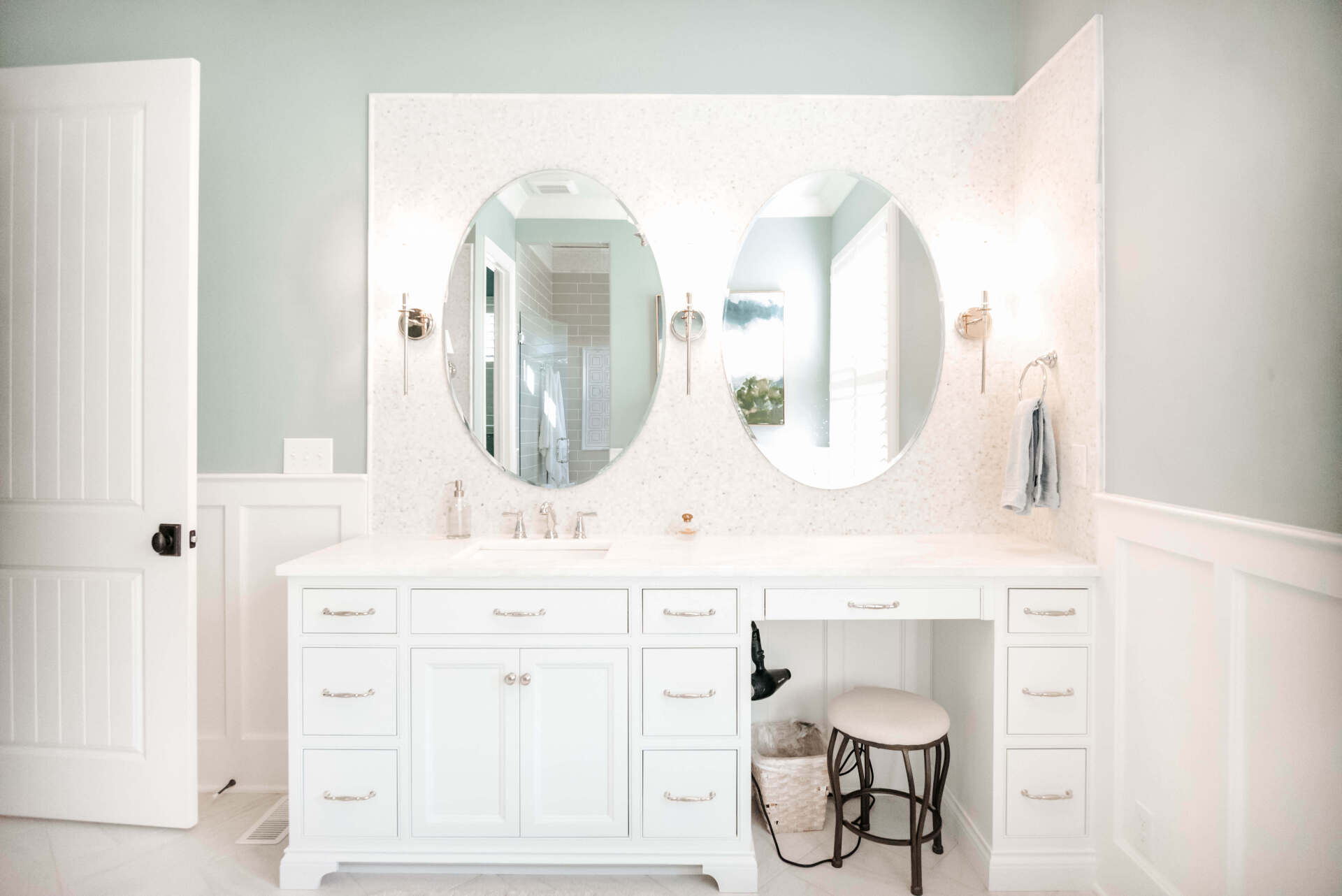 Bright white bathroom vanity with double oval mirrors and a small stool underneath
