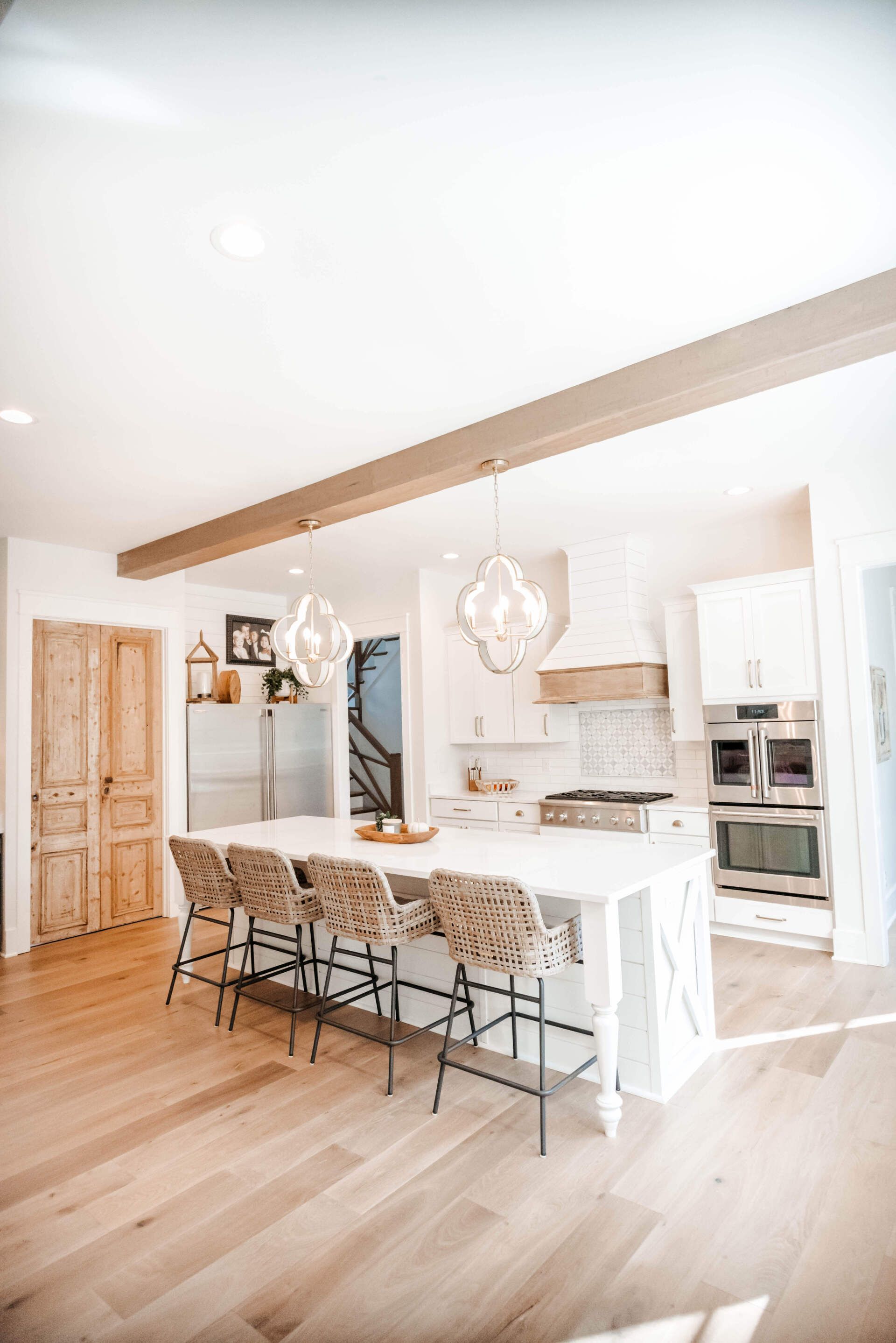 Bright white modern kitchen with island, woven stools, wood floor, and exposed ceiling beam