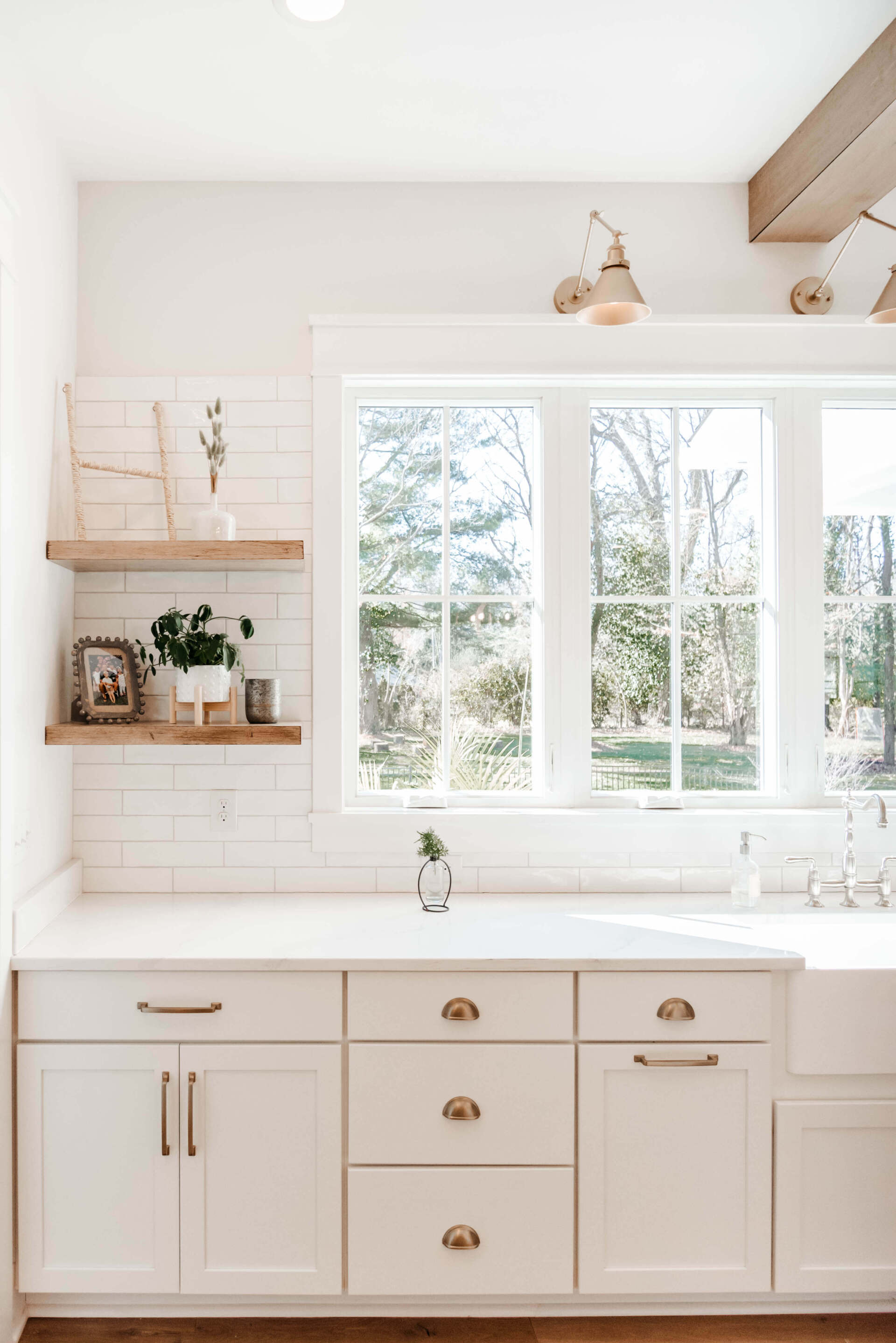 Bright white kitchen sink under large windows with open shelving and gold hardware