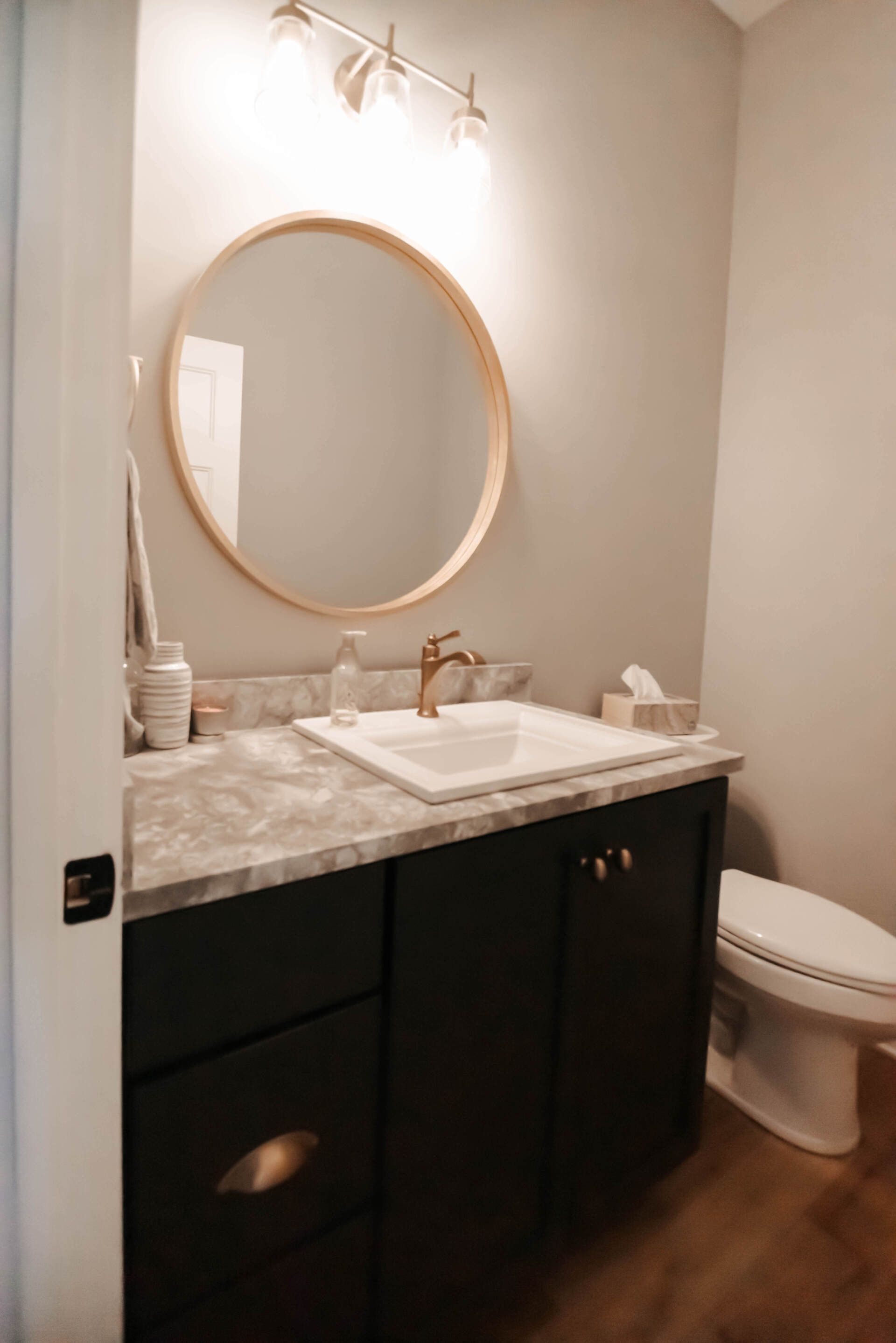 Small modern bathroom with dark vanity, round mirror, white sink, and toilet