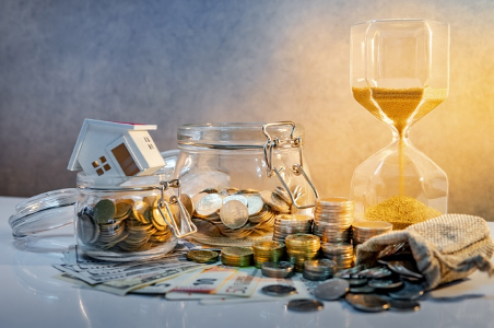 Glass jars of coins, cash, a house figurine, and an hourglass on a table, symbolizing savings and time.