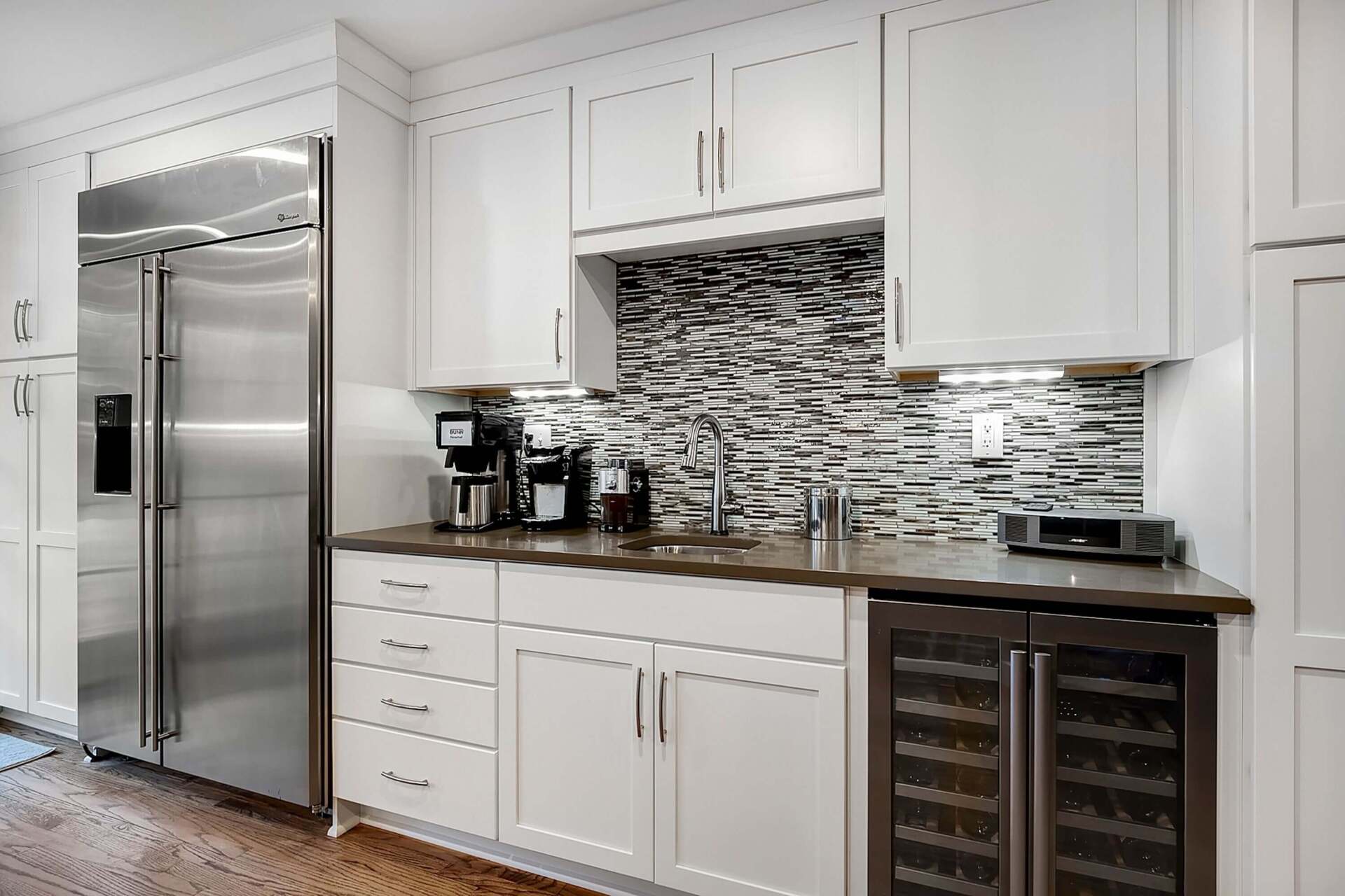 Modern white kitchen with stainless steel refrigerator, mosaic backsplash, coffee maker, and wine cooler