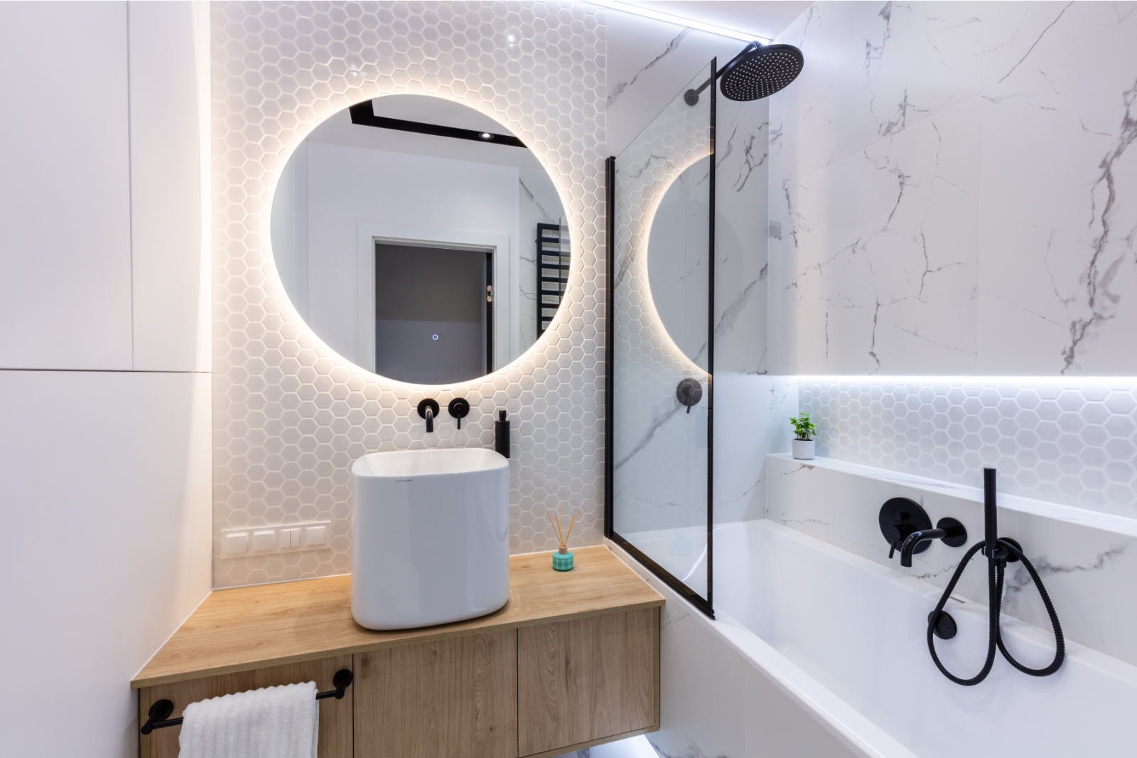 Modern bathroom with round lit mirror, wood vanity, black fixtures, and white marble tub surround