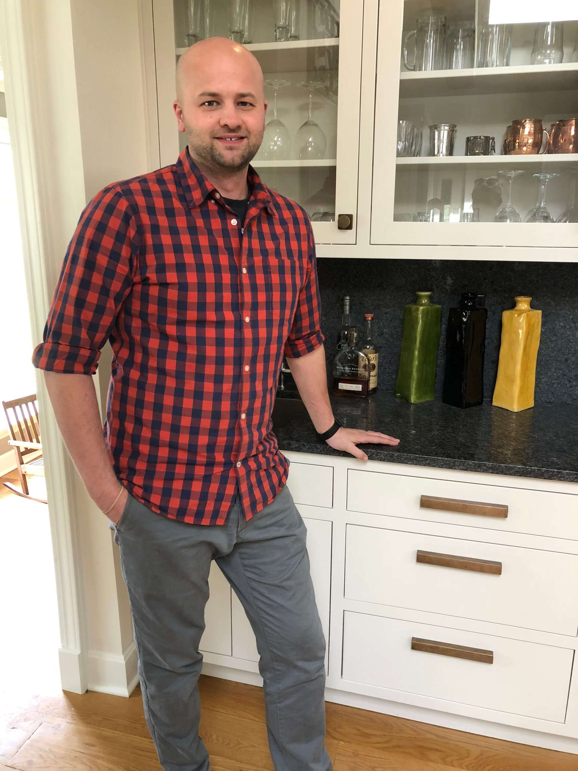 Man standing in a kitchen beside white cabinets, wearing a red plaid shirt and gray pants.