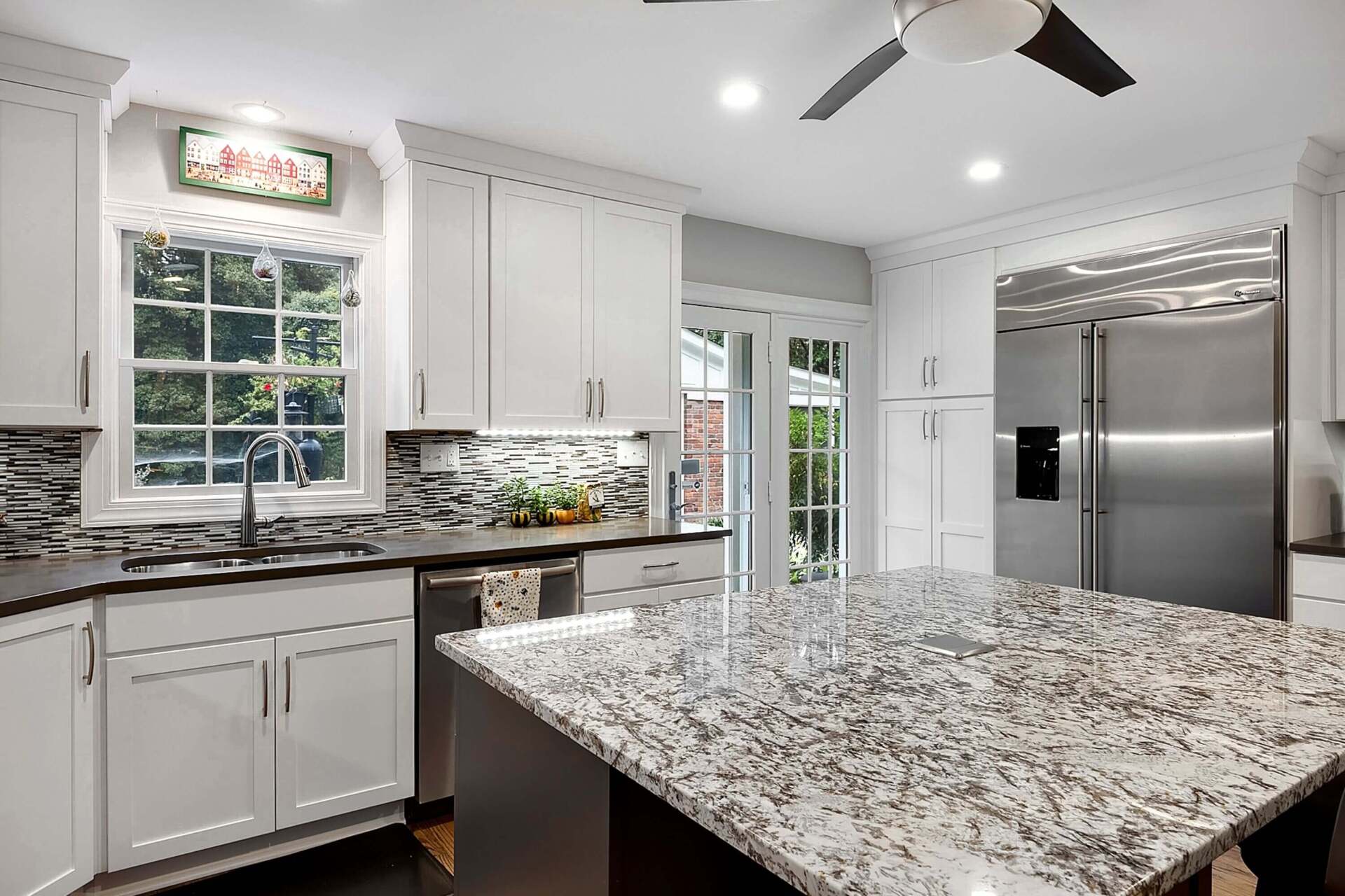Bright modern kitchen with white cabinets, granite island, stainless steel fridge, and large window