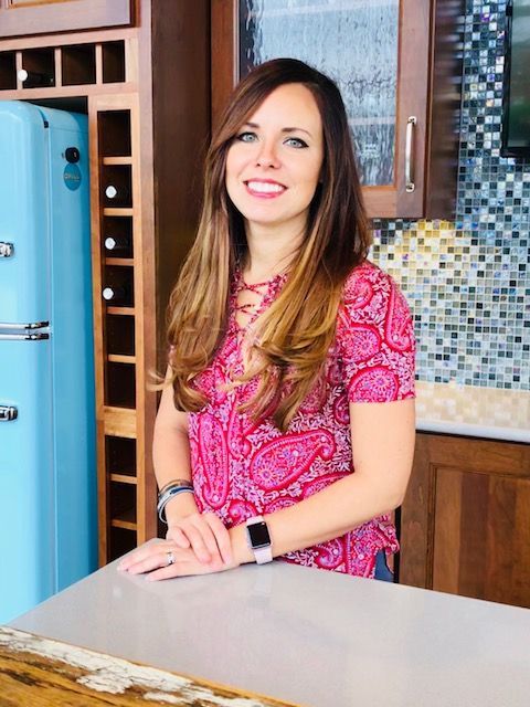 Woman smiling in a kitchen, wearing a pink patterned dress, standing by a countertop.