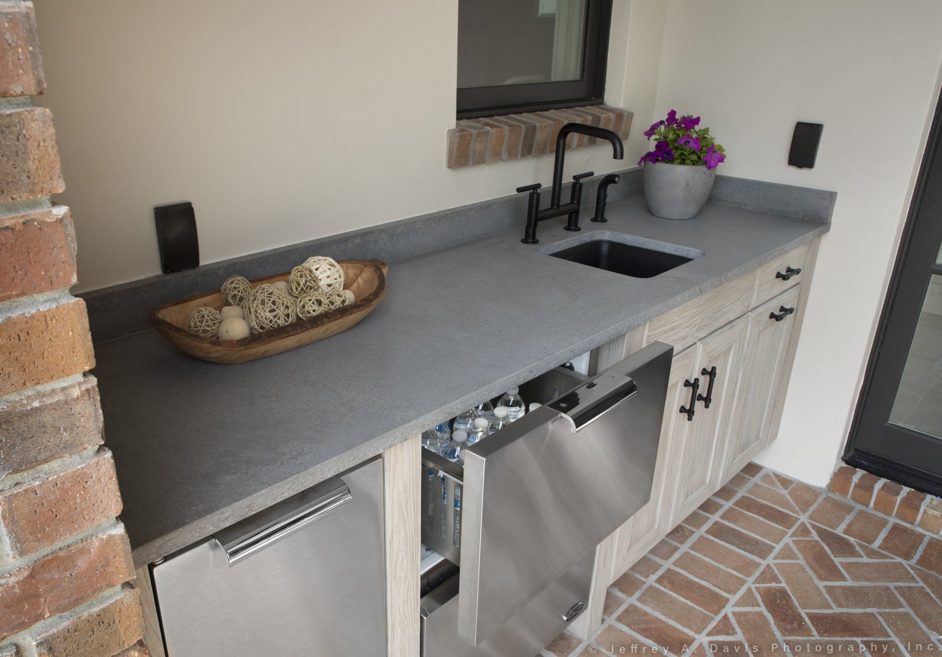 Outdoor stone countertop with sink, black faucet, basket, and potted flowers under a window