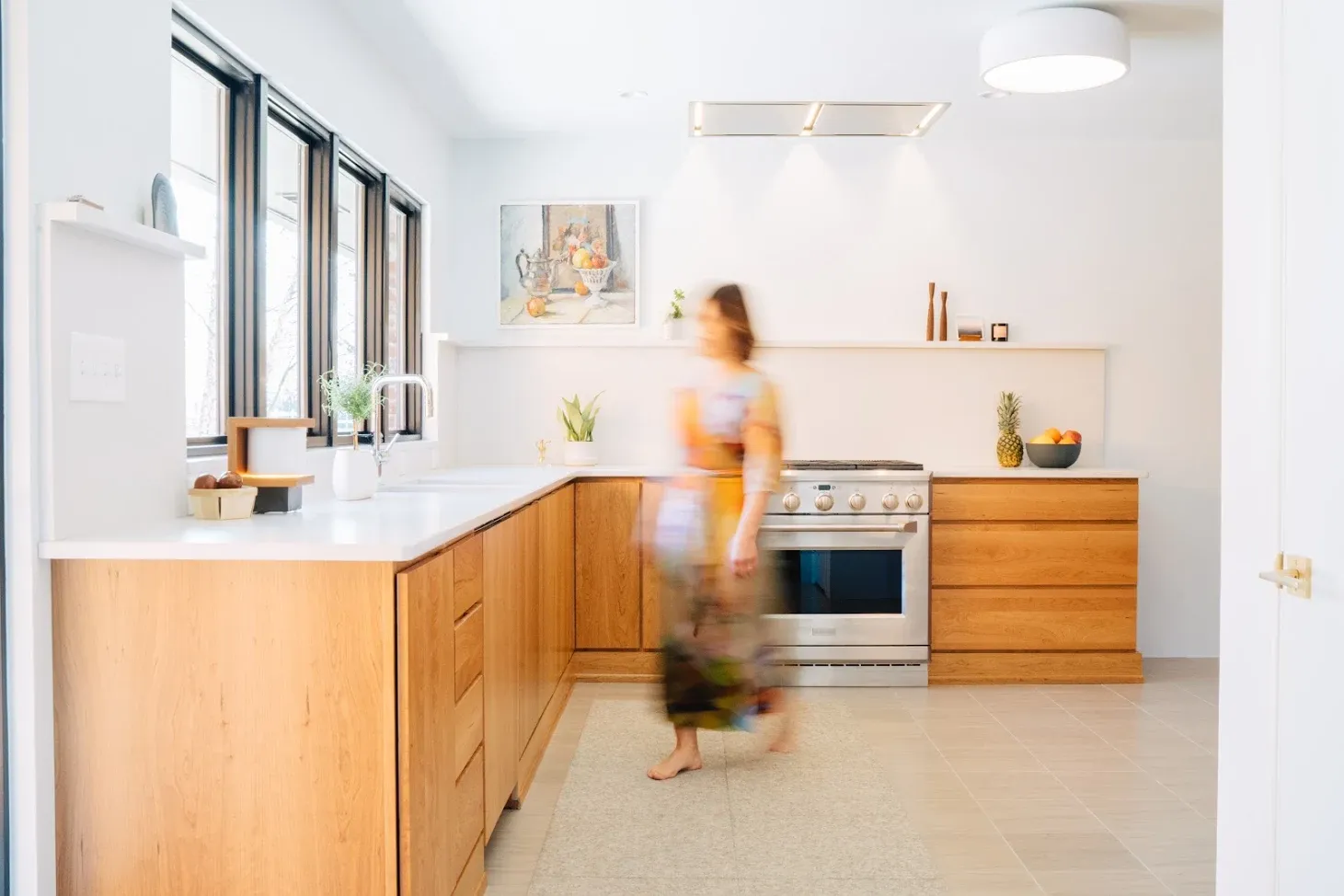 Bright kitchen with wooden cabinets, white countertops, and a blurred person walking near the stove.