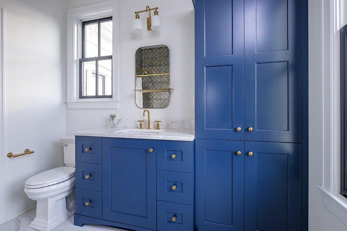 Blue bathroom vanity and cabinet with brass fixtures beside a white toilet and window