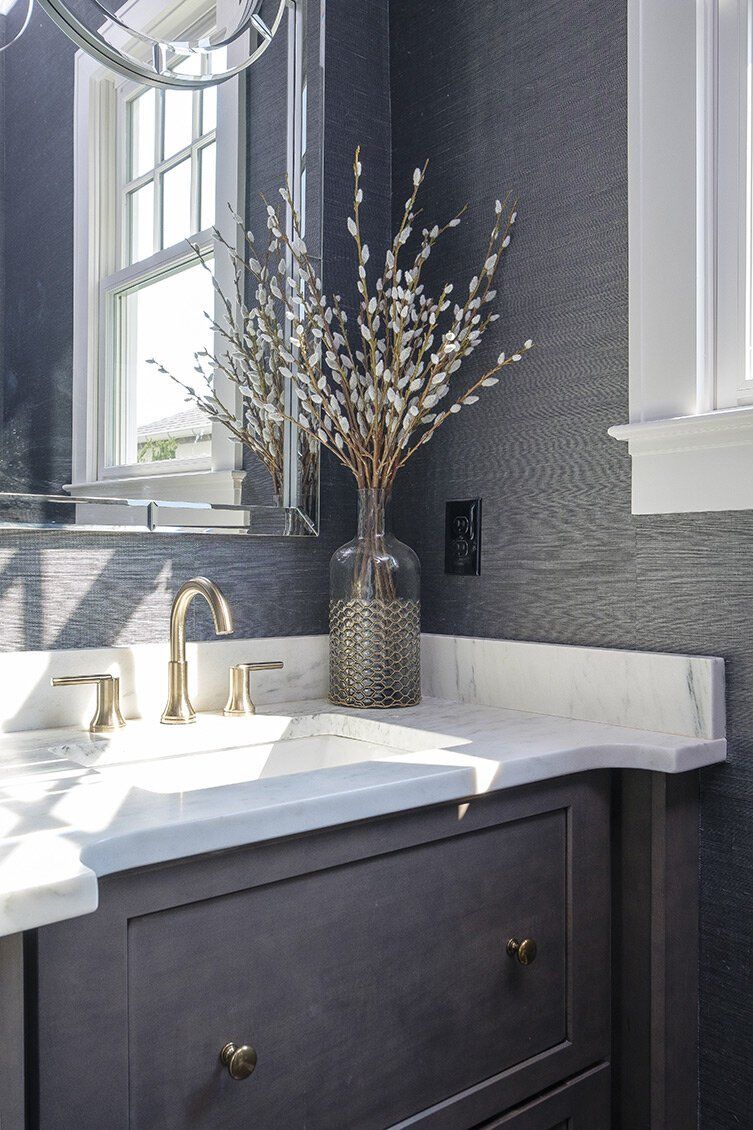 Dark vanity with white sink beneath a window, a vase of dried branches, and gray textured walls
