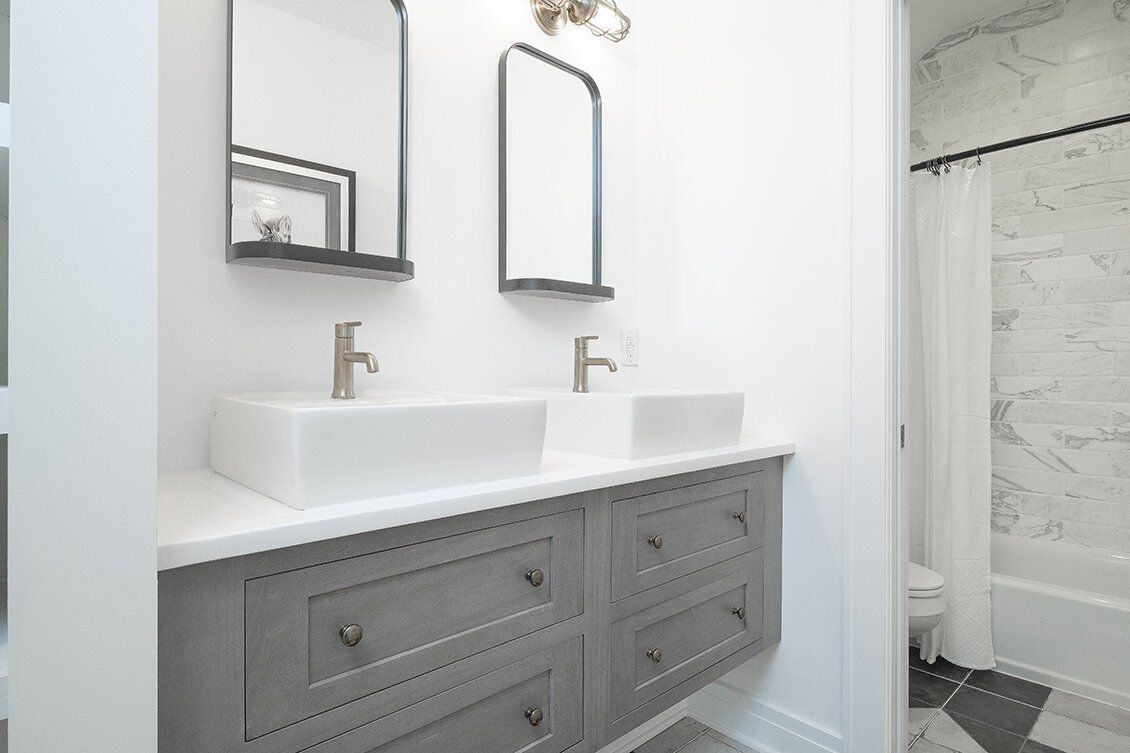 Bright bathroom vanity with double sinks, gray cabinets, mirrors, and a shower visible beside it