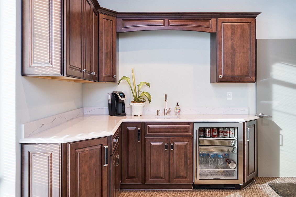 Small kitchen nook with wooden cabinets, white countertops, sink, and mini fridge under a window.