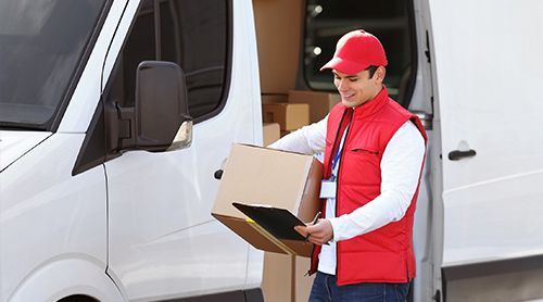 A delivery person in a red vest and cap holds a package and clipboard next to a white delivery van.