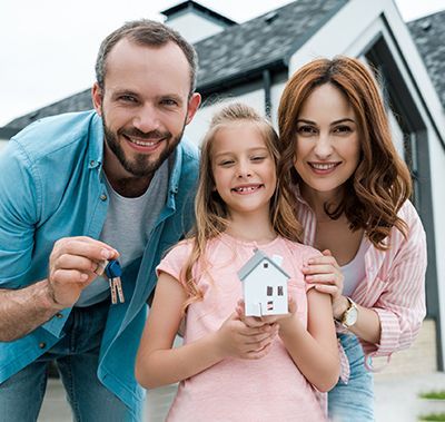 A smiling family stands in front of a new house, with a parent holding keys and a child holding a model of a home.