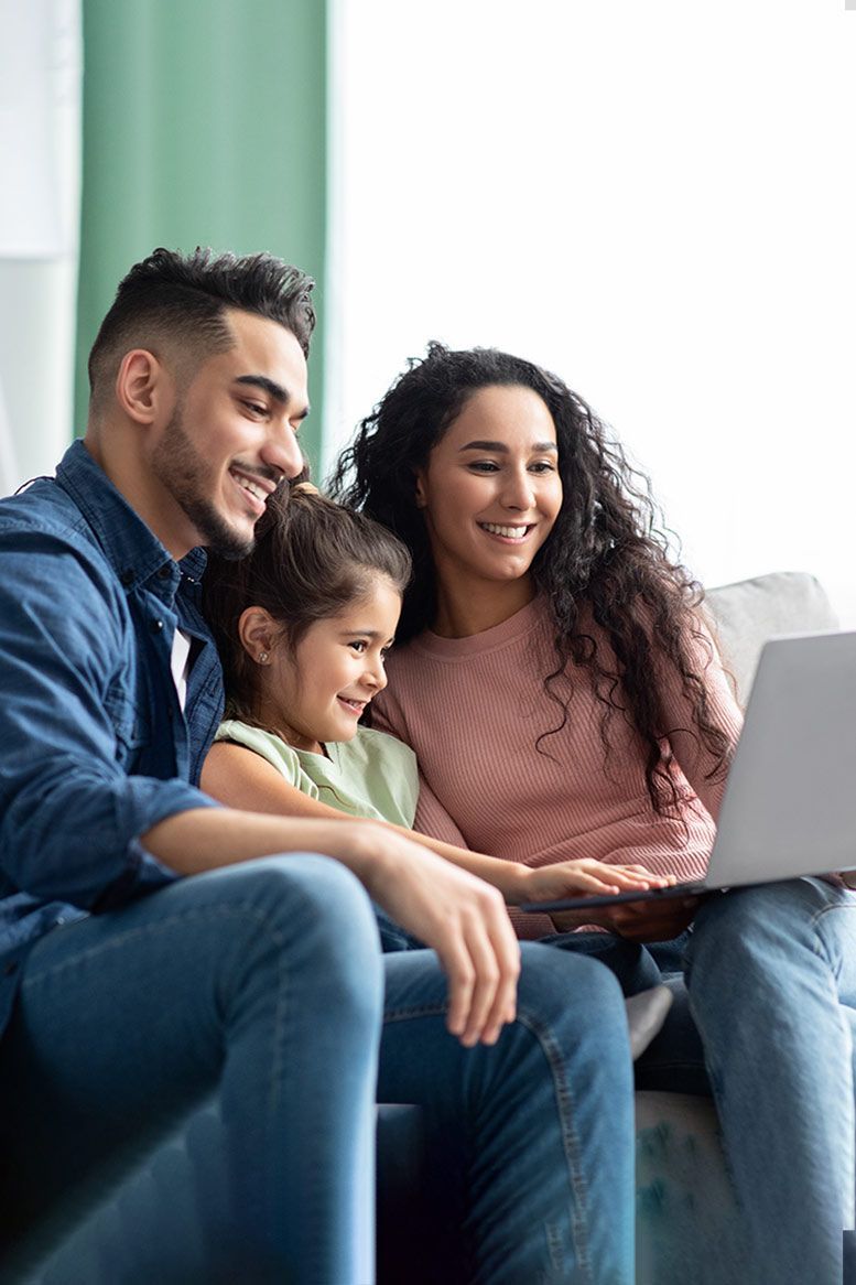 A family sits together on a couch, smiling while looking at a laptop computer.