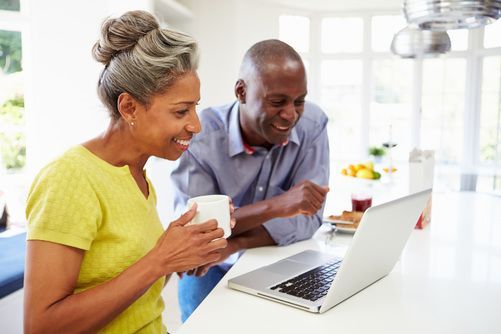 Two people sitting at a white kitchen counter, smiling and looking at a laptop together while holding a white mug.