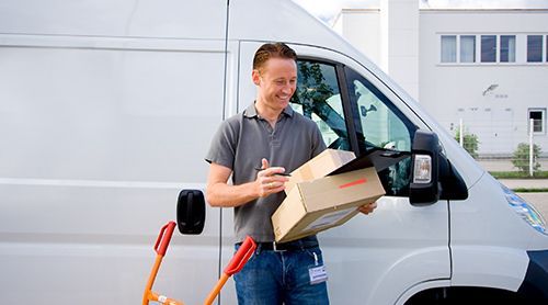 A delivery worker in a grey polo shirt smiling while holding a cardboard box in front of a white delivery van.