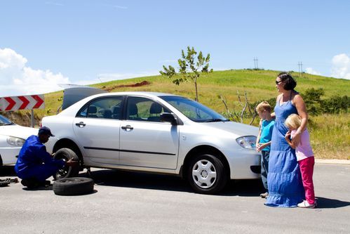 A mechanic changes a tire on a silver sedan on the side of a road, while a woman and two children stand nearby watching.