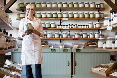 A person in a white lab coat holds a jar in a shop filled with shelves of labeled glass spice or herb jars.