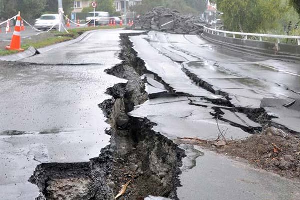 A large fissure splits a paved road, causing the asphalt to buckle and break under a gray, overcast sky.