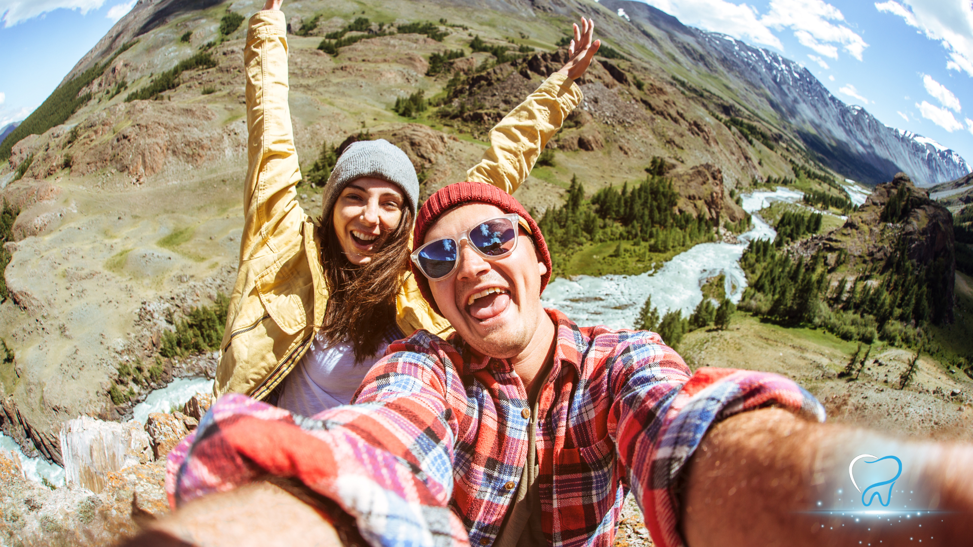 Two smiling hikers take a selfie with mountain peaks and a river valley behind them.