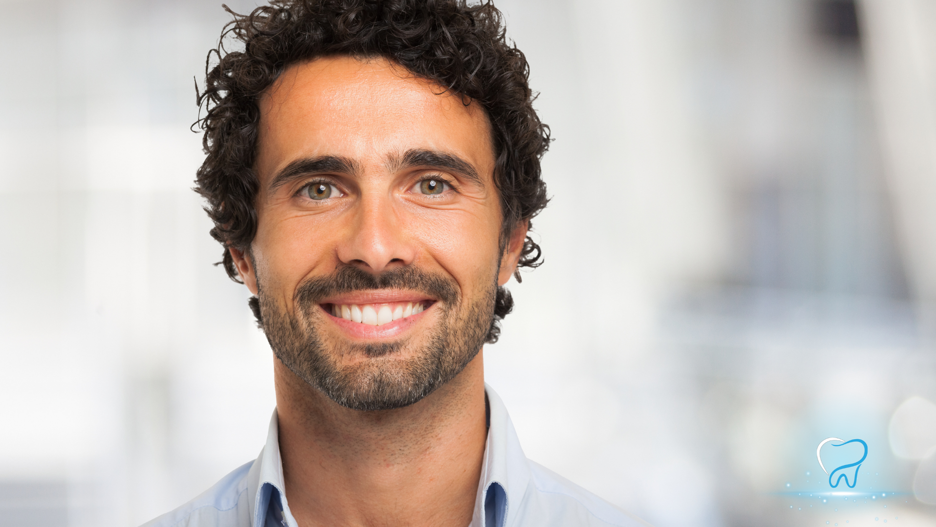 Smiling man with curly dark hair and beard against a blurred light background