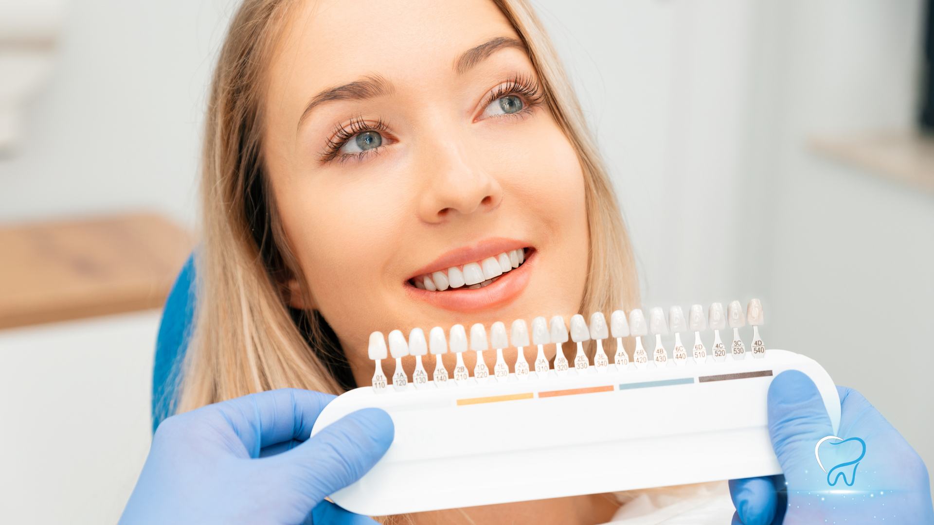 Gloved hands hold a dental shade guide near a smiling woman in a clinic
