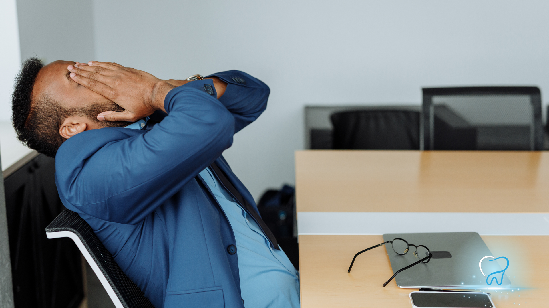 Man in blue shirt leaning back at a desk, hands over face, looking stressed in an office
