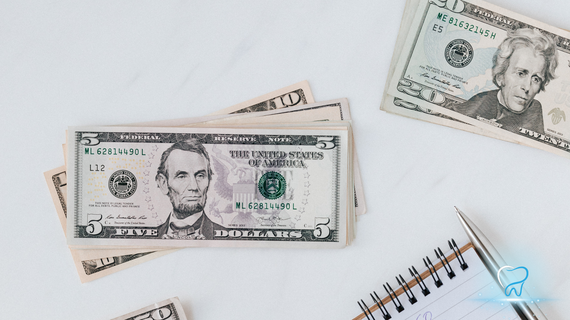 Scattered U.S. dollar bills on a white surface next to a small spiral notebook and blue paperclip