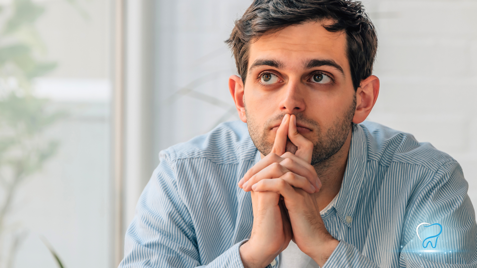 Thoughtful person in a light blue shirt resting clasped hands near their face indoors.