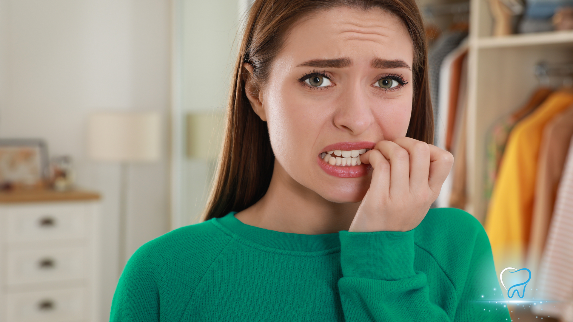 Young woman in green sweater biting her nails, looking worried in a bright room with clothes behind her