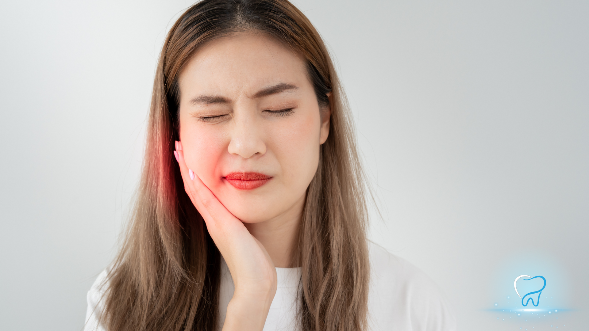 Woman with eyes closed, holding her cheek in pain against a plain light background.