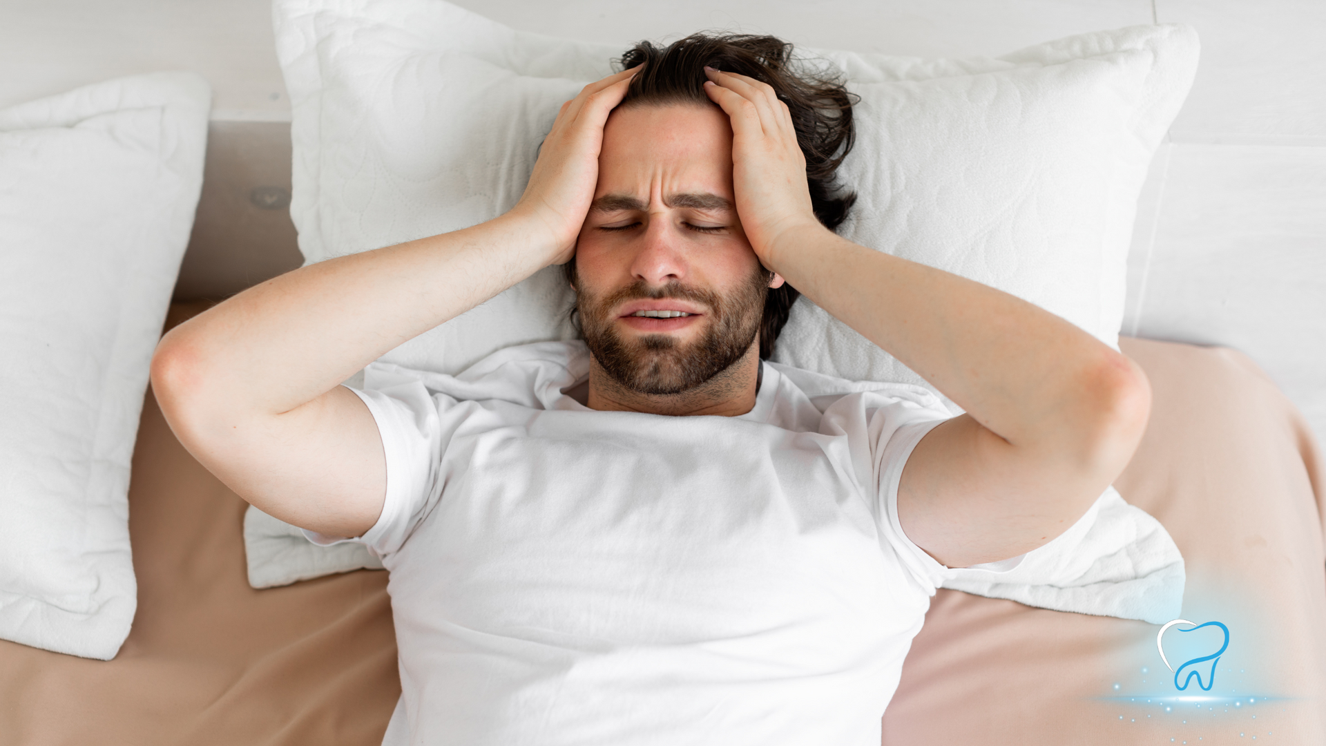 Man lying in bed holding his head, looking stressed or in pain, on white pillows and sheets