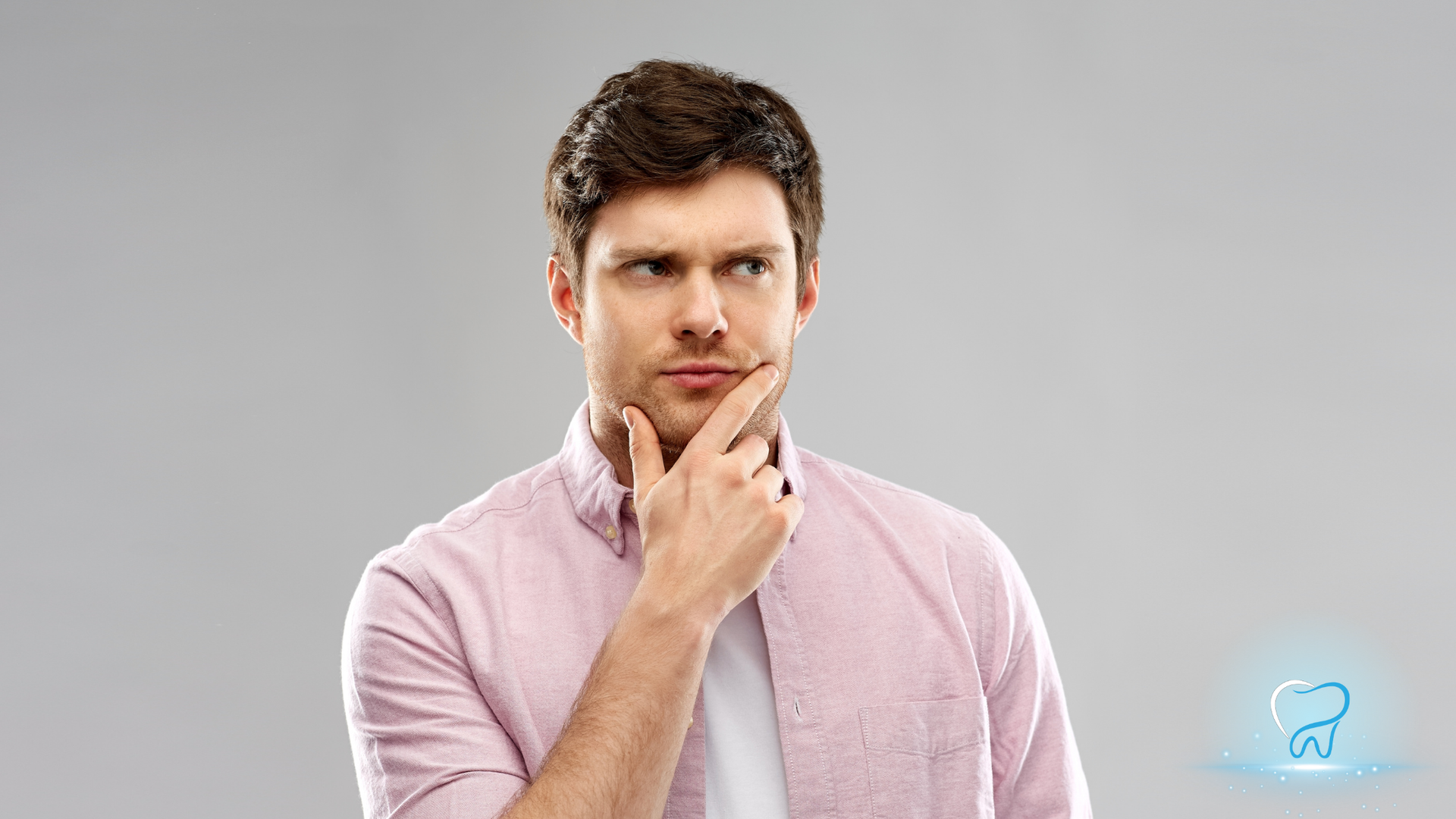 Man in a pink shirt and tie, touching his chin with a thoughtful expression against a gray background.