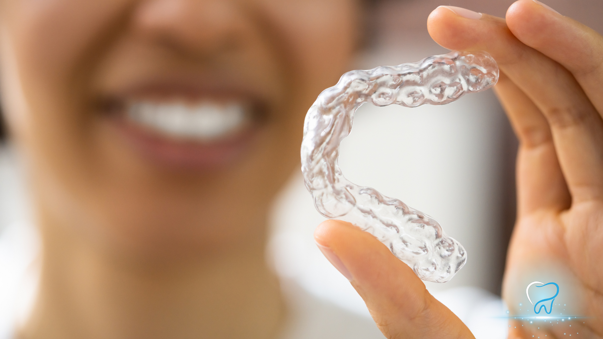 Hand holding a clear plastic dental aligner in front of a smiling mouth