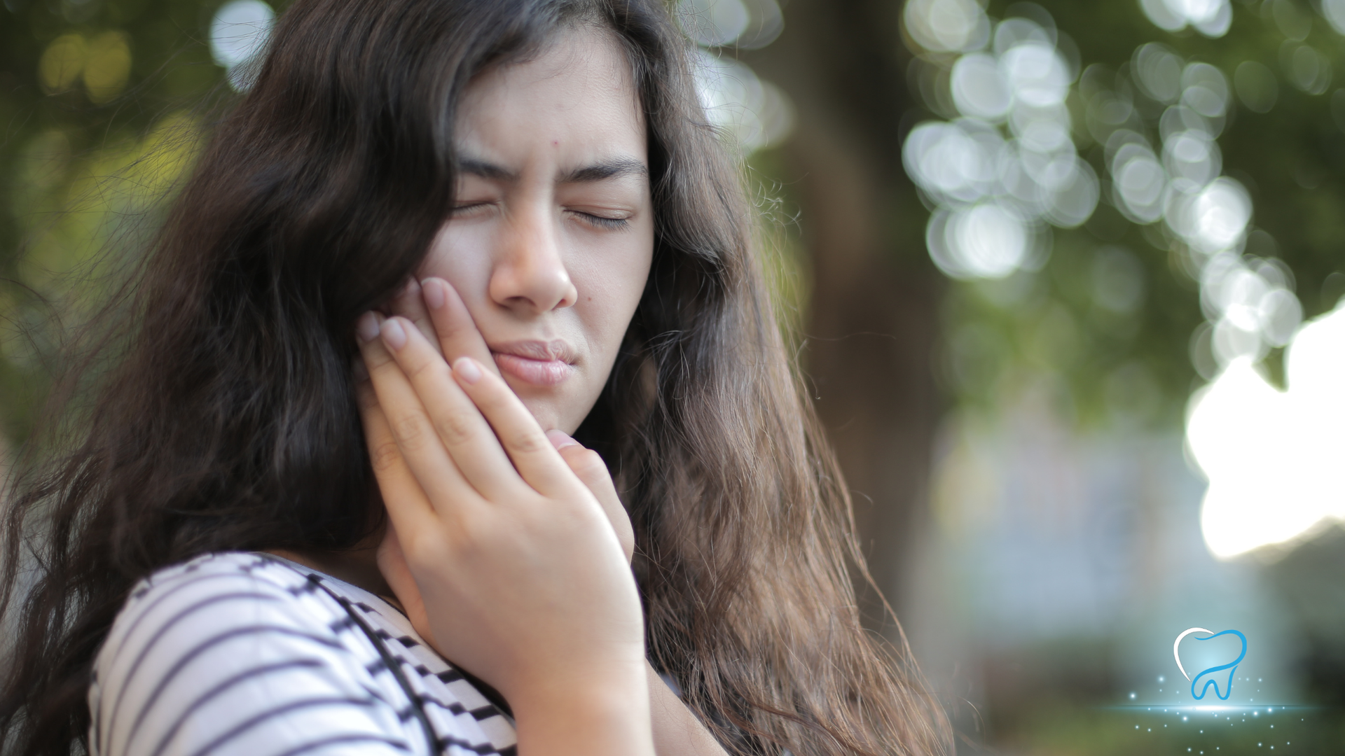 Woman holding her cheek with eyes closed, appearing to be in pain outdoors near blurred trees.