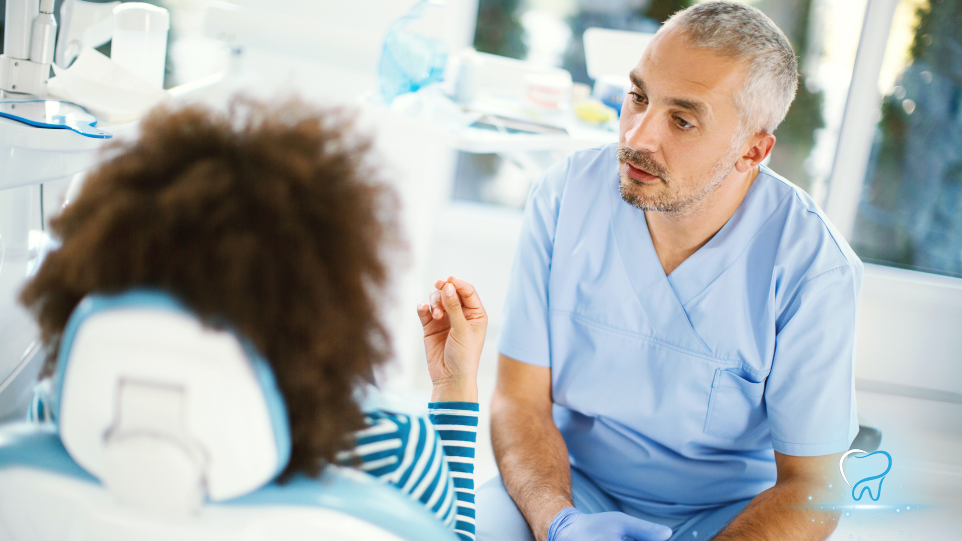 Dental professional in blue scrubs speaking with a patient in a bright clinic, holding a small instrument.