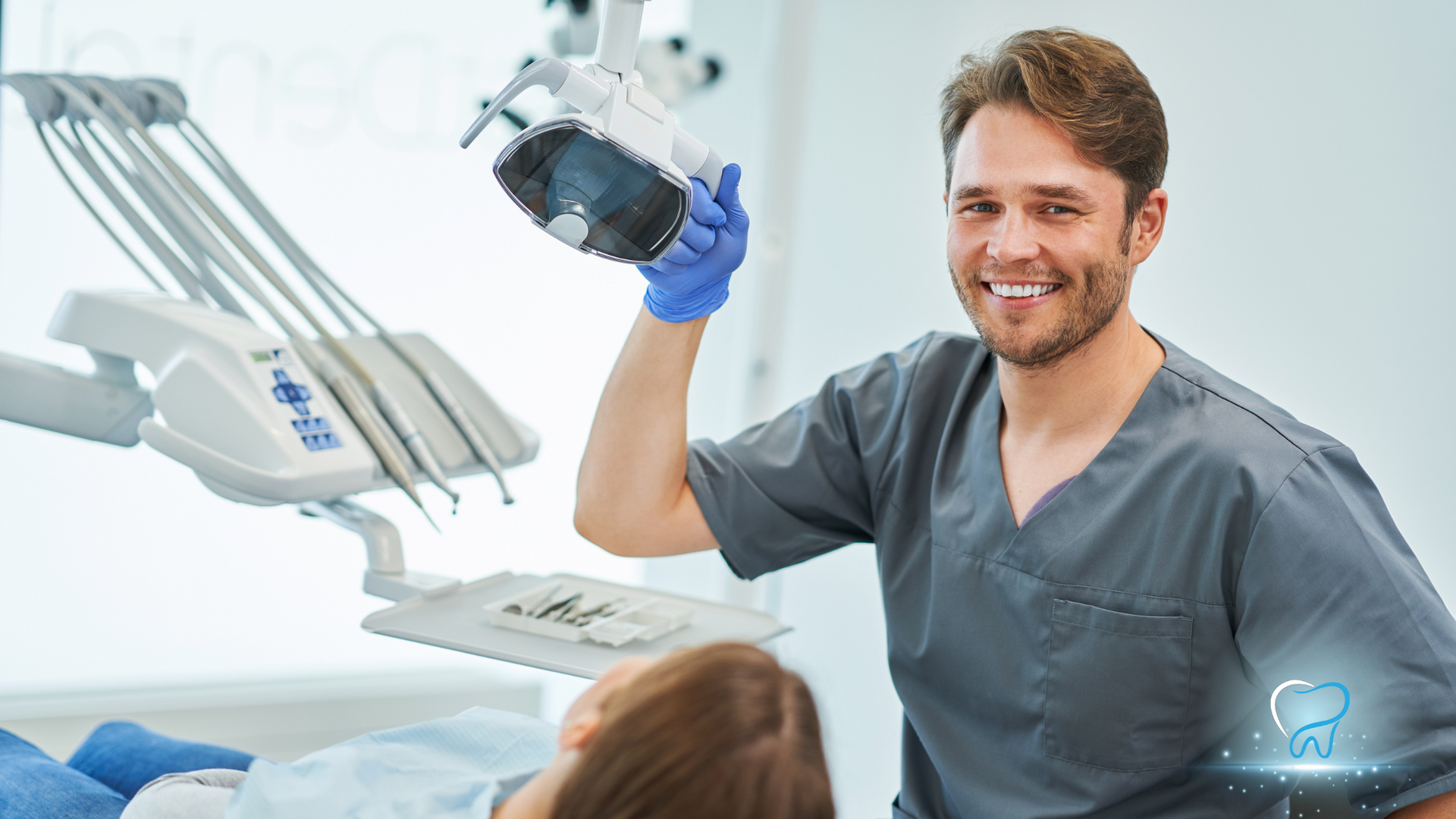 Dentist smiling in a dental clinic, holding a black dental tool near a reclining patient.