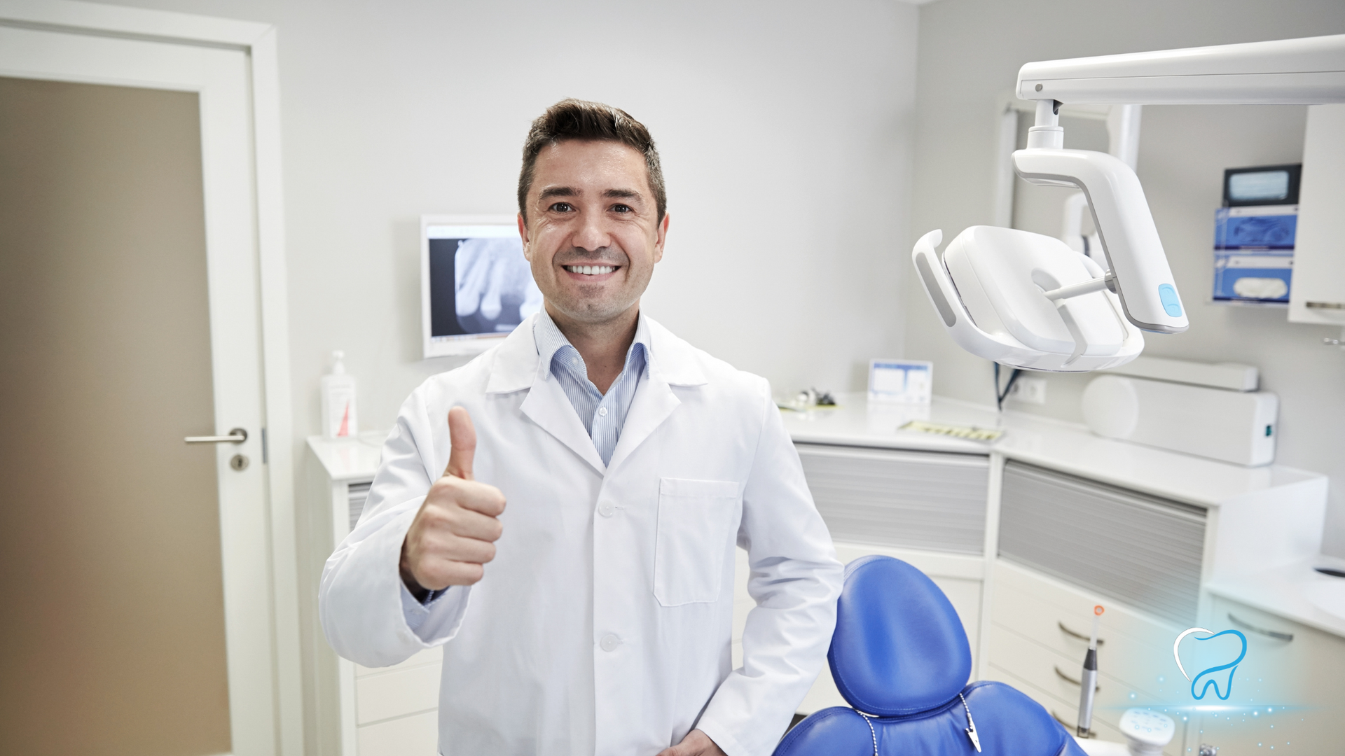 Smiling dentist in white coat giving a thumbs-up in a bright dental clinic beside a blue chair